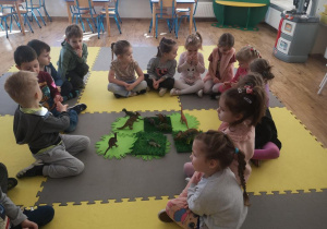 Widok na siedzące w kręgu na macie dzieci, które przekazują sobie kawałek węgiel i soli. W środku kręgu widać plastikowe dinozaury ułożone na zielonych serwetkach i trawkach.